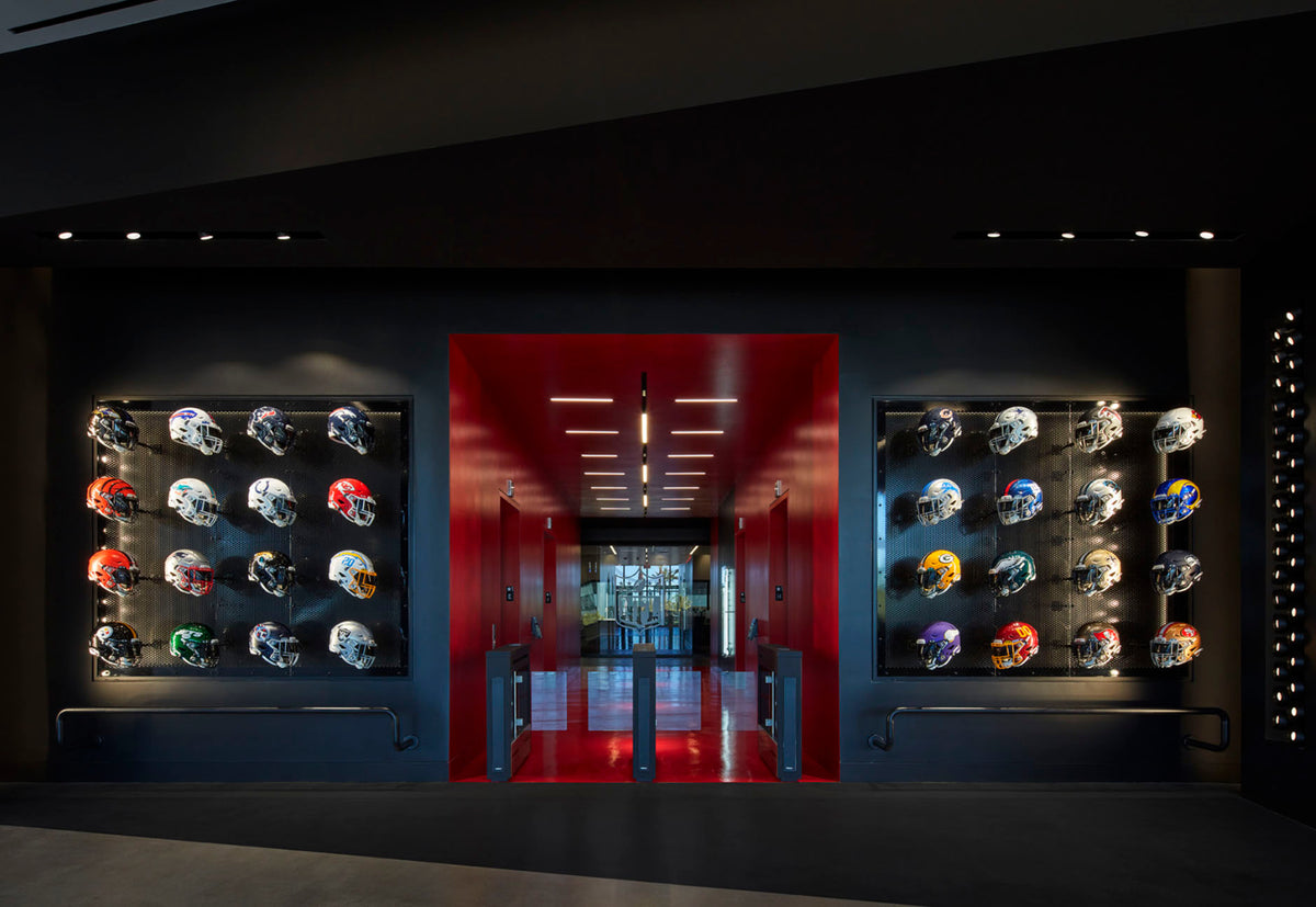 A long, dark interior hallway framed by two illuminated wall displays of football helmets in various colors, with a vivid red doorway in the center. Accent lighting.
