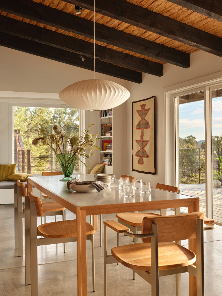 A modern dining room featuring a light wood dining table with matching wooden chairs, a large white pleated pendant light, and a high ceiling with dark wood beams and a woven reed ceiling panel. EL Design