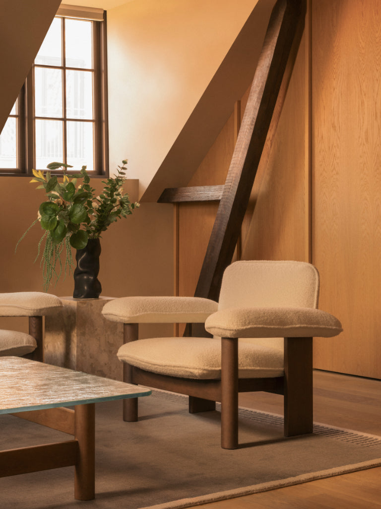 A mid-century modern armchair with cream bouclé upholstery and dark wood legs, styled in a warm, sunlit room with wooden walls, a slanted window, and a coffee table. EL Design