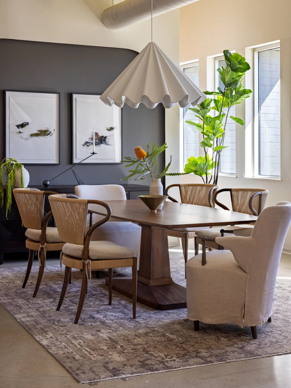 A dining room scene with a rectangular wooden dining table surrounded by upholstered chairs, including a prominent beige armchair in the foreground. The room has a dark gray accent wall with framed artwork, a large white pleated pendant light, tall windows, and a potted green plant.