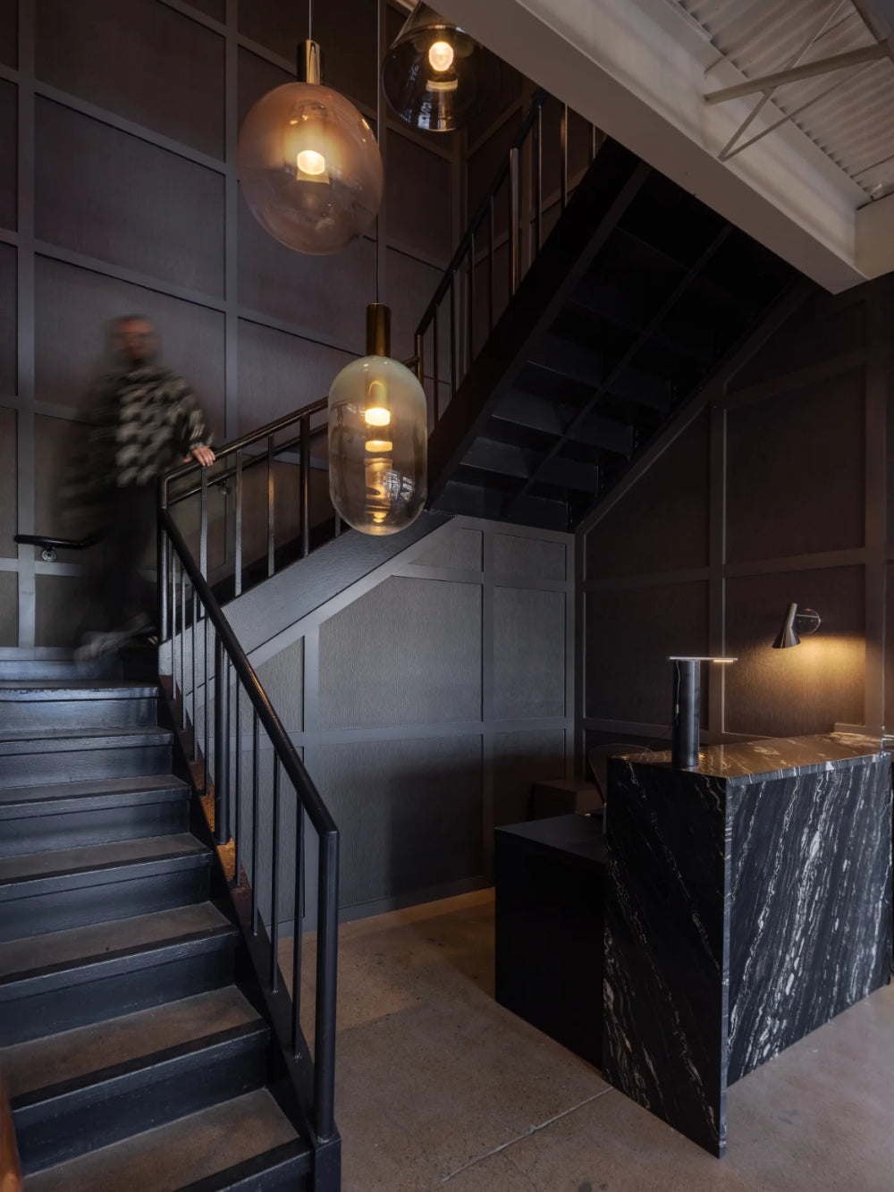 A modern interior scene with a dark staircase, pendant lights in glass shades, a marble reception desk, and a blurred person walking along the stairs.