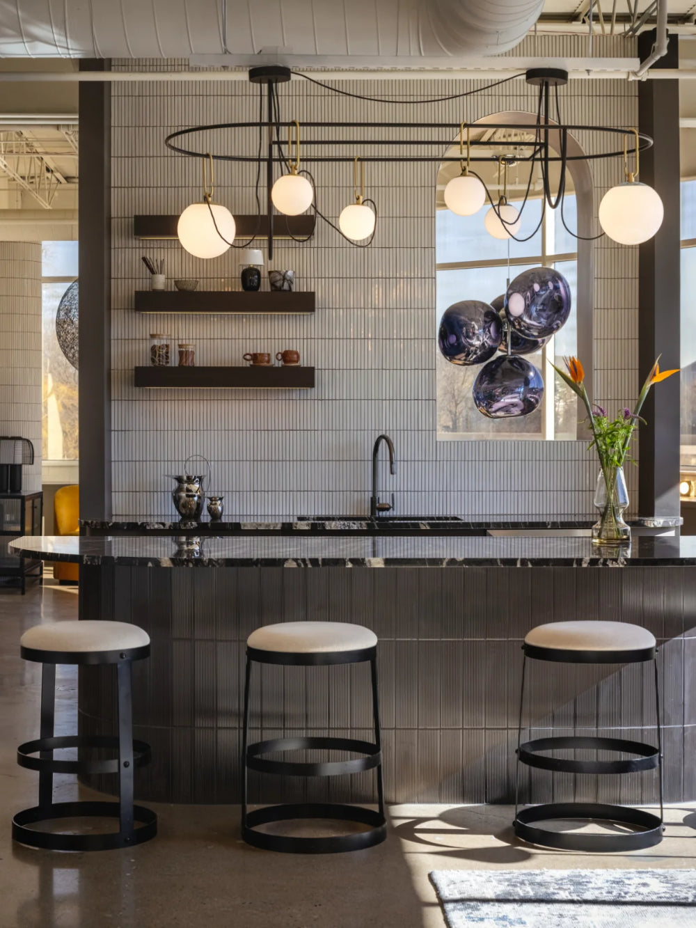 A modern kitchen island with a dark wood countertop and three black bar stools with cream cushions, set in a contemporary kitchen with white subway tiles, floating shelves, and a large window.