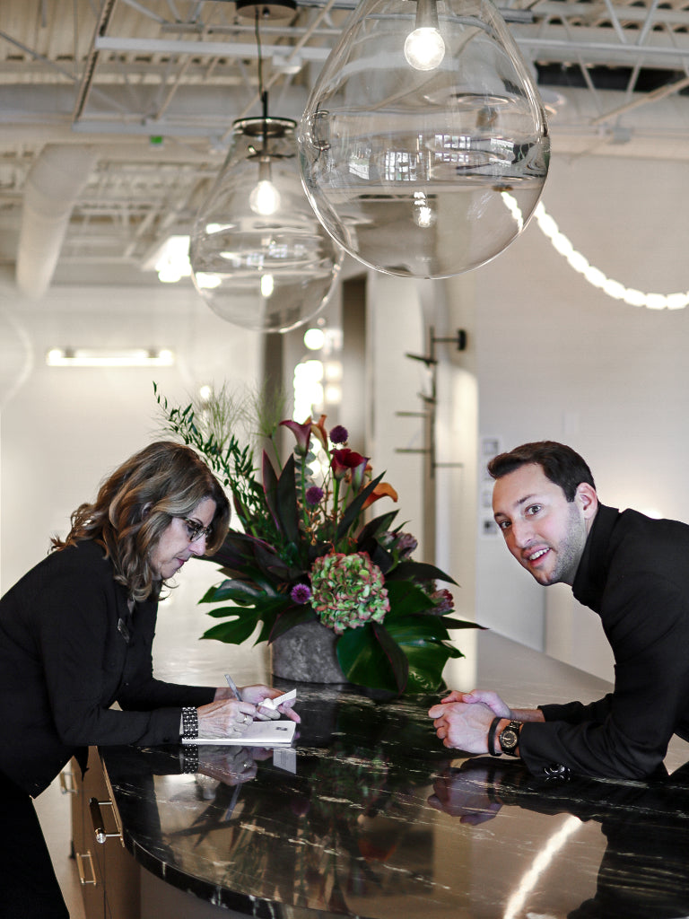 Two people sitting at a table with a large floral arrangement in the center.