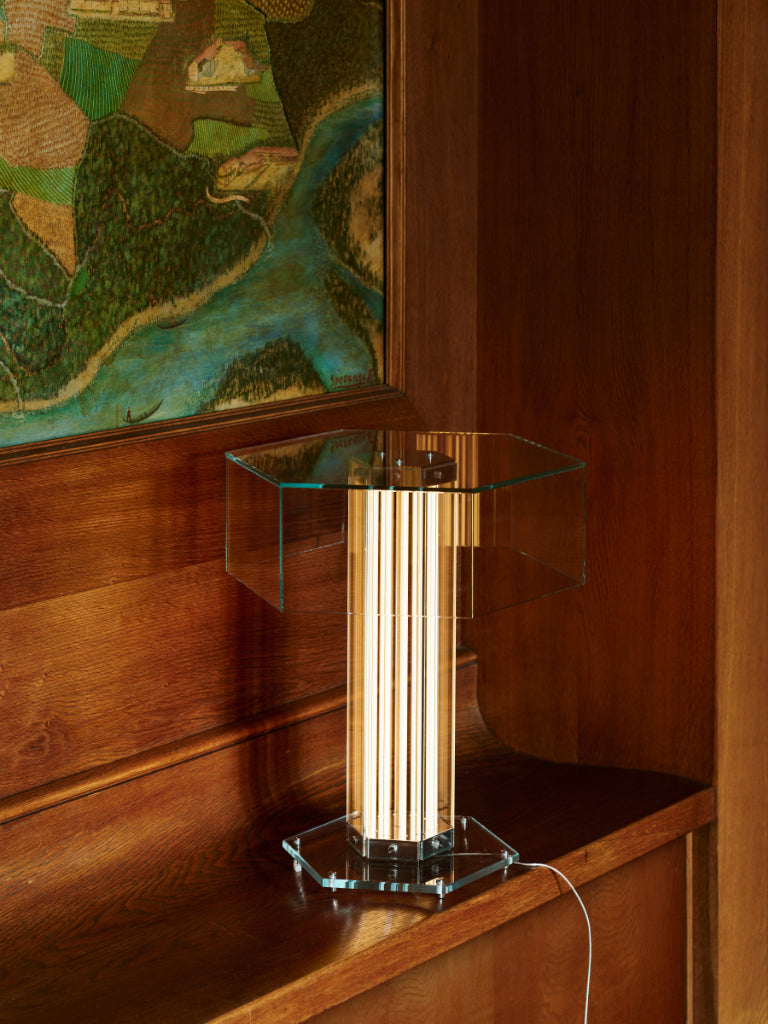 A modern table lamp with a hexagonal clear glass top and base, featuring a central column of vertical light-wood slats glowing warm white, set on a wooden console next to a framed landscape painting. EL Design