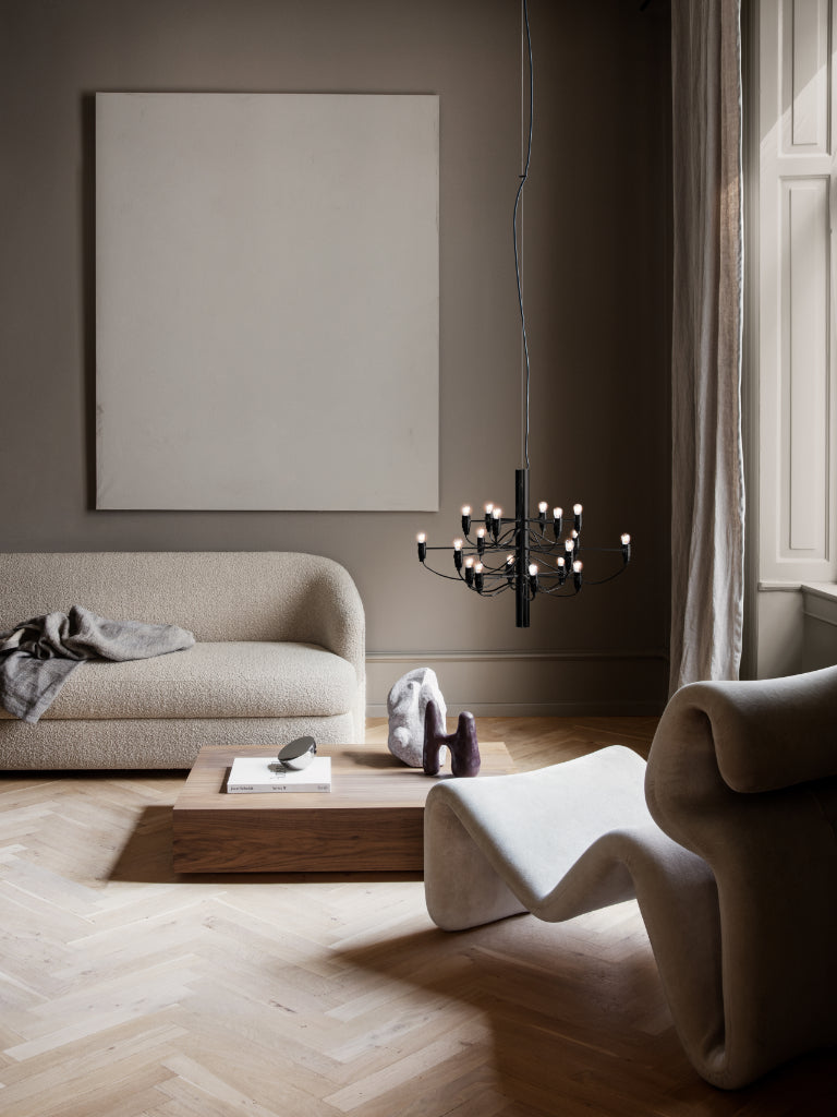 A modern living room scene with a beige textured sofa, a low wooden coffee table, sculptural white seating, a black multi-light chandelier, and a large blank canvas painting on a dark taupe wall. EL Design