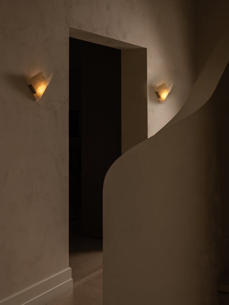 Two modern wedge-shaped wall sconces with warm glowing bulbs mounted on textured beige walls, flanking a dark doorway in a minimalist interior setting. EL Design