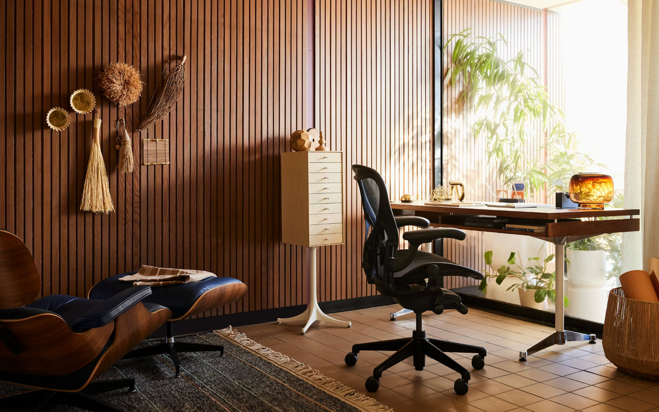 A modern home office interior with a brown wood slat wall, black ergonomic office chair, desk, sideboard, plants, and a brown leather lounge chair designed by Herman Miller.