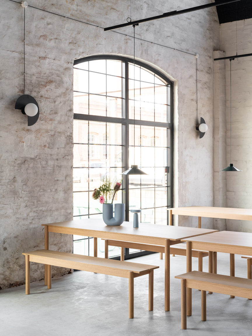 Modern dining area with wooden tables and benches against a concrete wall with large windows.