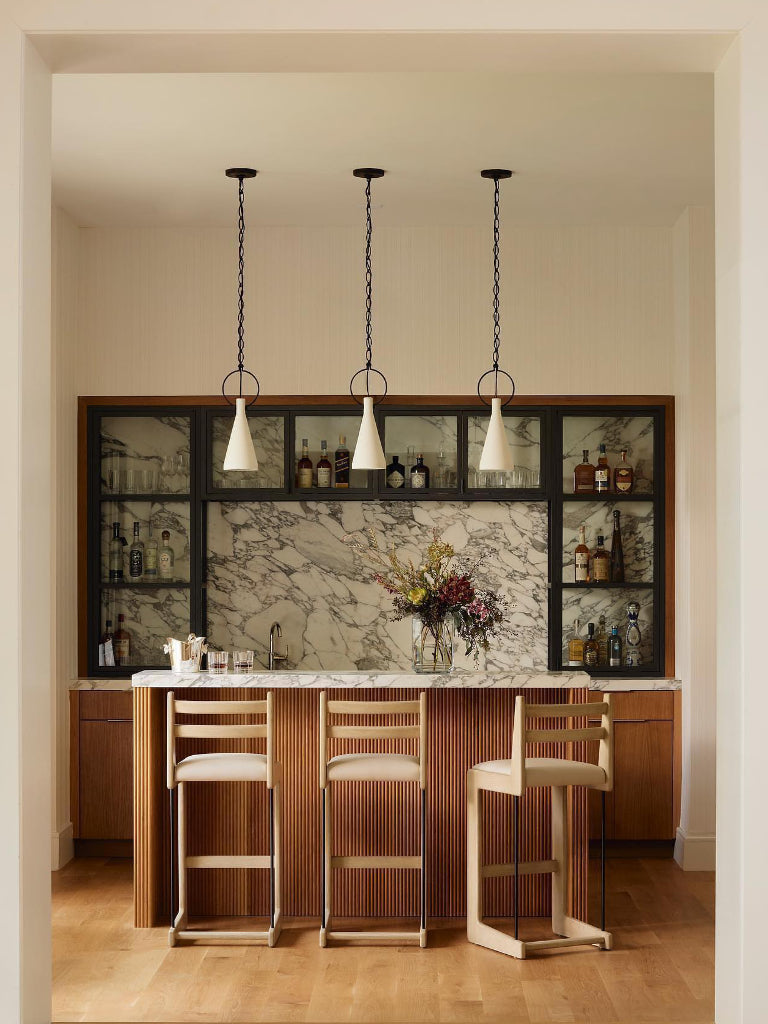 A modern bar area with a marble-patterned backsplash, dark wood shelving, a slatted wood bar front, three hanging pendant lights, and three bar stools in a warm interior setting. EL Design