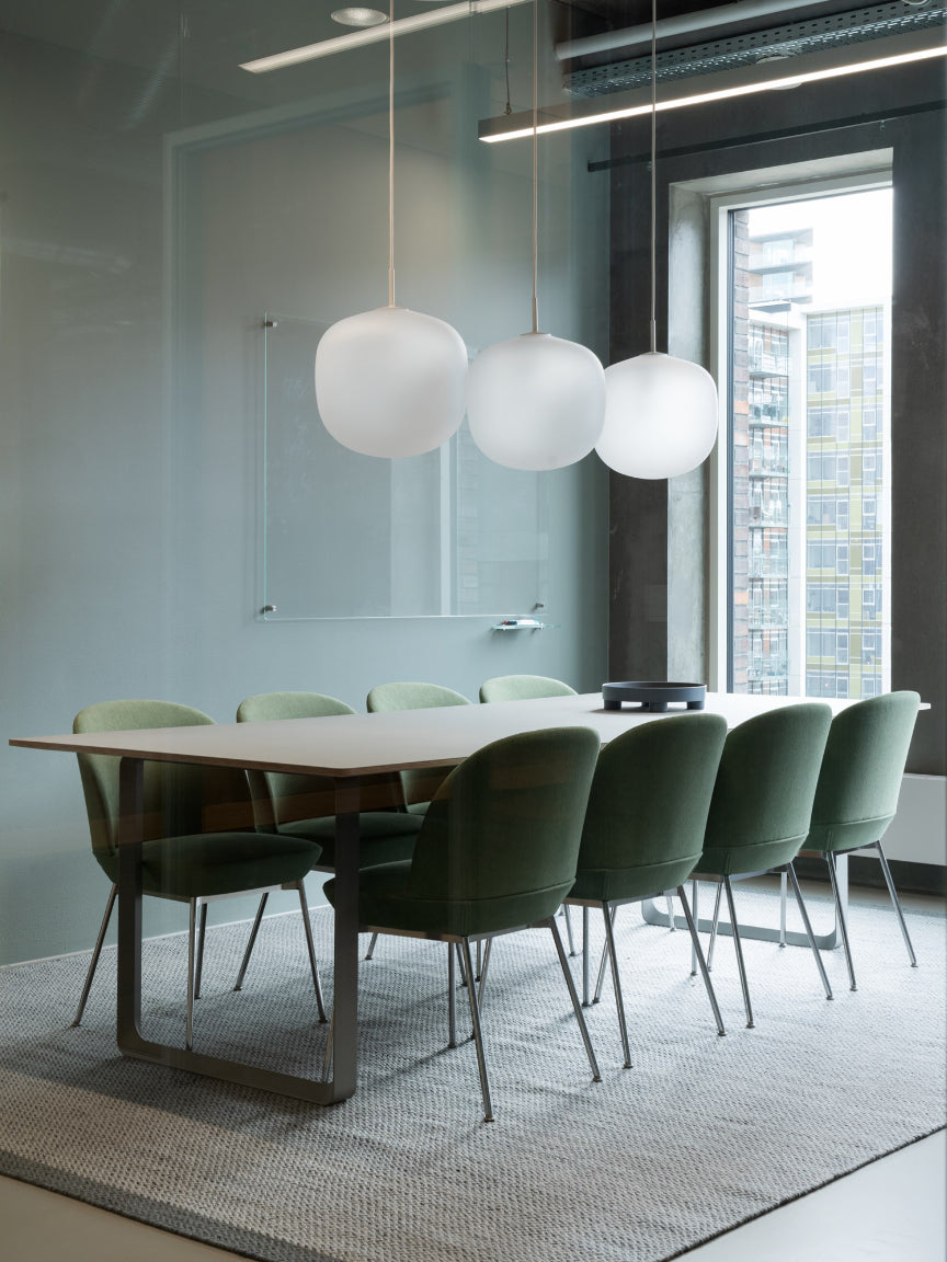 Modern conference room with a long wooden table and green chairs, featuring pendant lights and large windows.