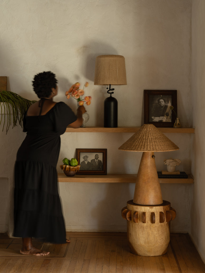 Woman arranging flowers on a shelf with decorative lamps and framed pictures in a warm-toned room.