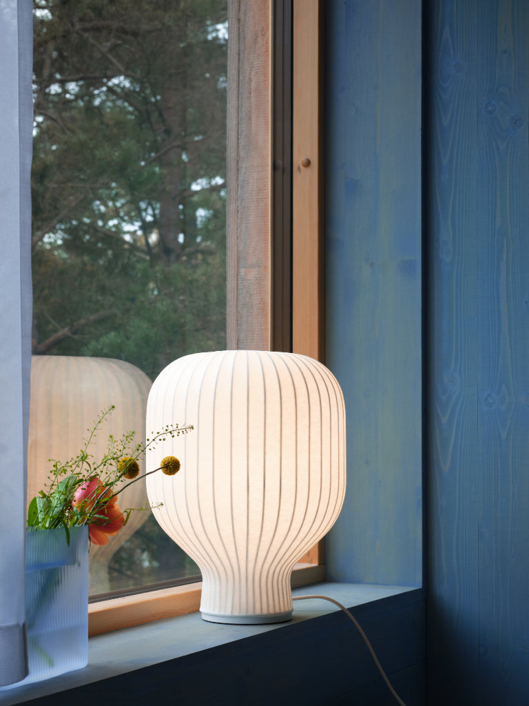 White ribbed table lamp on a windowsill with a view of trees outside.