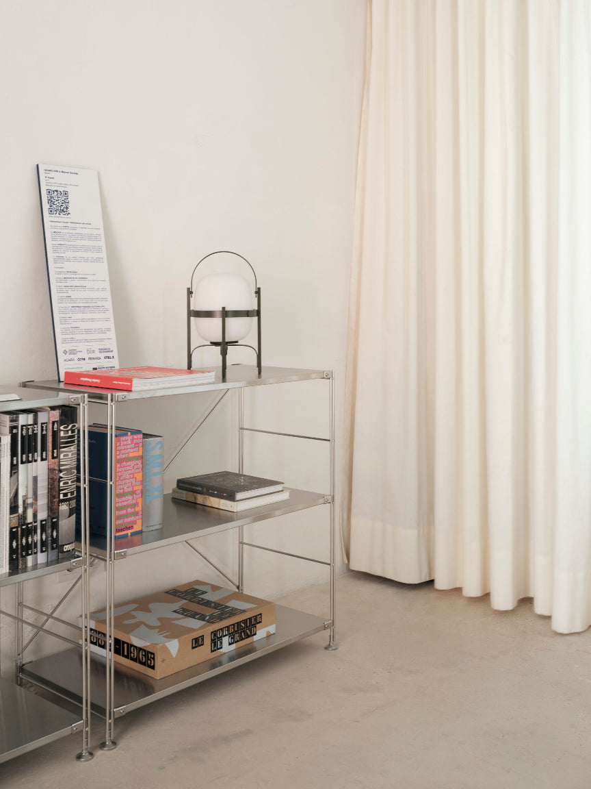 Metal shelving unit with books and a lamp against a white wall with a curtain.