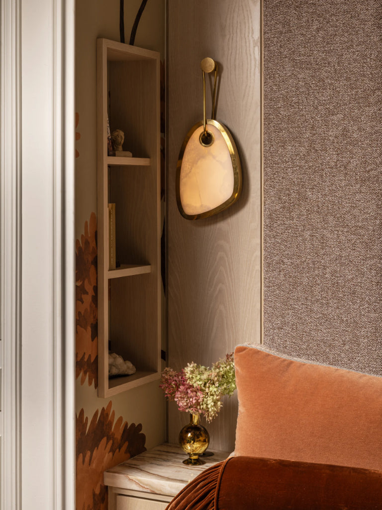 Decorative wall light fixture on a textured wall with a shelf and flowers in the background.
